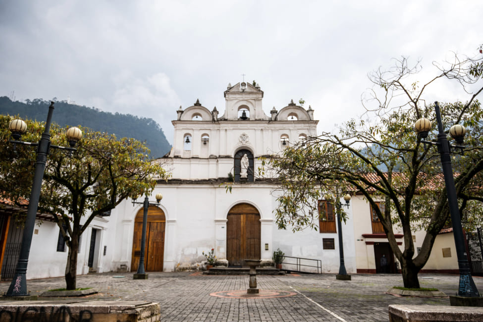 Parroquia de Nuestra Señora de Las Aguas fachada frente completa blancacon dos puertas gigantes en madera, dos ventanas, campanario con palomas y de fondo los cerros orientales. Ramas de árboles a los costados.