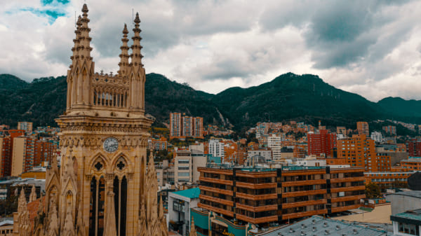  Iglesia de Lourdes foto de dron de la parte alta en la que se ve el reloj y las agujas,, y el cielo cubierto de nubes blancas. Está rodeada de edificios comerciales y al fondo se observan los cerros orientales con algunos edificios.