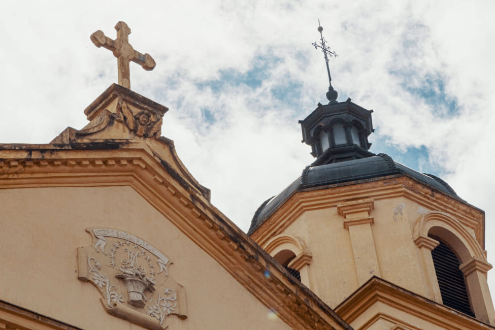 Iglesia de La Candelaria, parte superior de una de las torres de color entre amarillo y negro terminada en cúpula con cruz metálica negra mirando al cielo azul con nubes blancas.