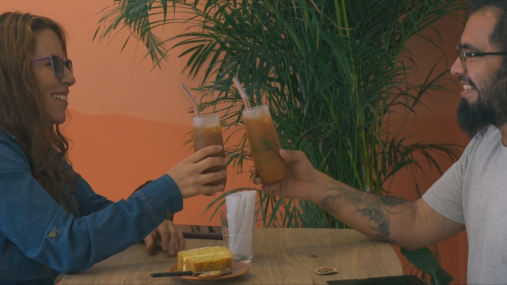 Un hombre y una mujer joven brindan con dos copas que parece contienen jugo. Rodean una mesa en la que hay un pedazo de torta y una cucharita.