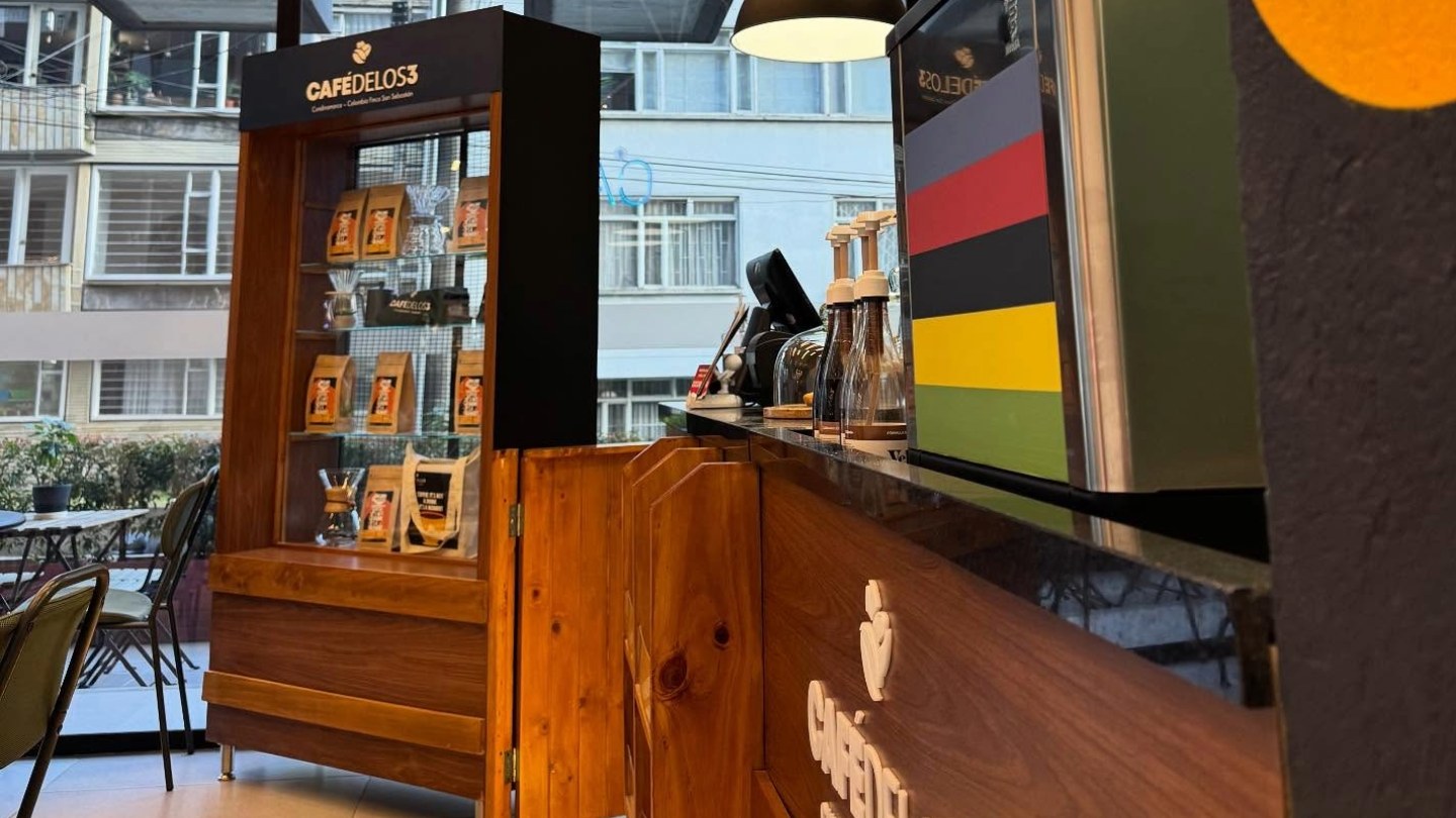  Interior de un local de día en el que se ve a un lado una barra con accesorios para hacer café, un anaquel de madera con café en bolsas. Hay unas mesas con sillas alrededor y al fondo un gran ventanal hacia la calle donde se ve la fachada de un edificio.