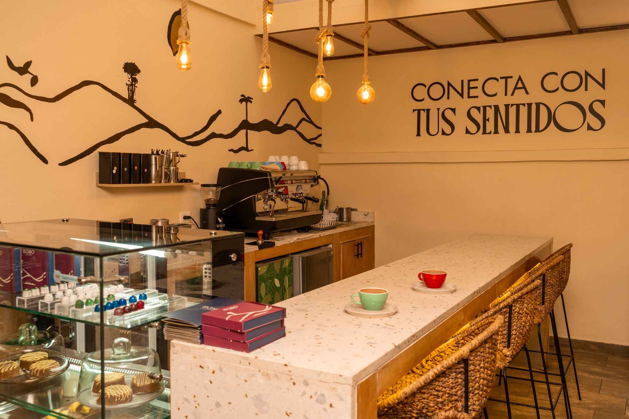 Interior de un local comercial cuyas paredes tienen color es castaño claro y en donde hay una vitrina con chocolates y tortas, y una grán máquina para preparar café. 