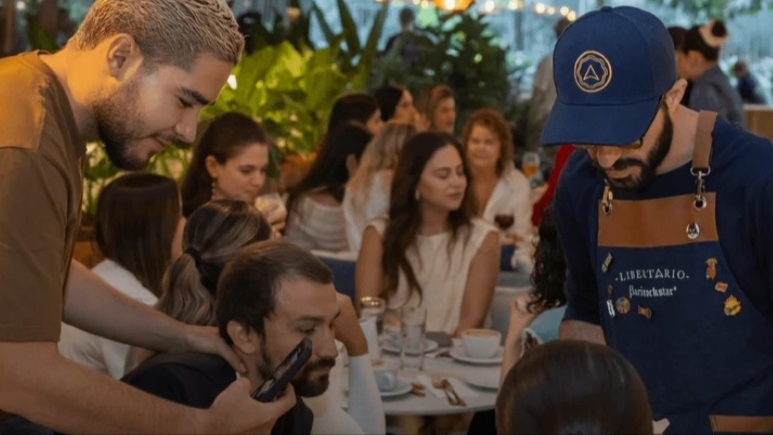 Grupo de personas jóvenes en un restaurante escucha a un mesero jóven que viste un delantal azul.
