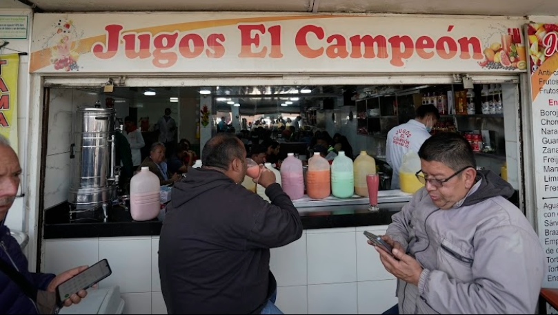  Fachada de un local comercial en donde hay un aviso que dice "Jugos El Campeón". Hay varios hombres adultos al frente y al fondo varias personas.