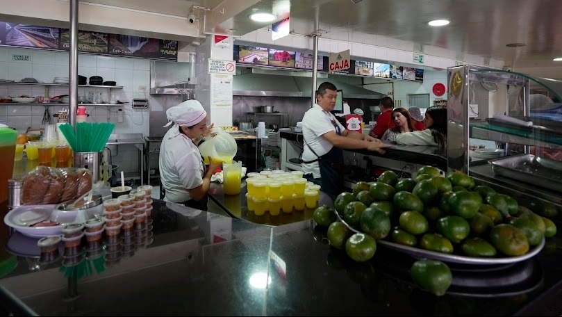 Interior de una amplia cocina en el que un hombre y una mujer adultos están preparando jugos. Se ven varias mandarinas verdes.