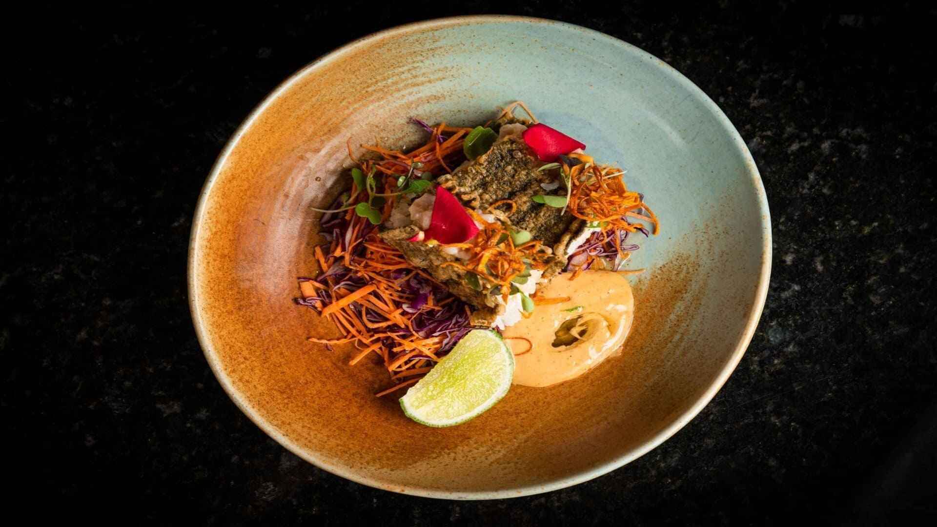 Un plato gourmet de pescado frito con ensalada de col, aderezo cremoso y una rodaja de lima en un cuenco artesanal.