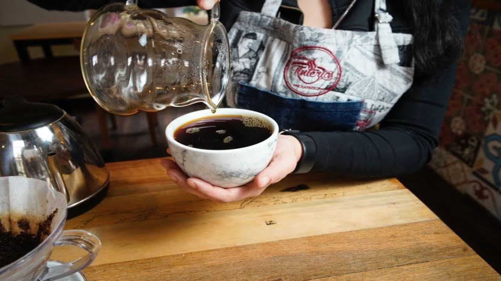 Una barista sirviendo café de goteo en una taza de mármol, con un delantal temático de café.