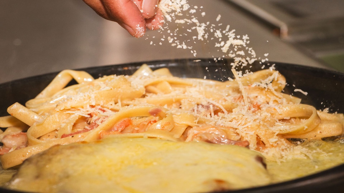 Una mano espolvorea generosamente queso rallado sobre un plato de pasta cremosa en una sartén.