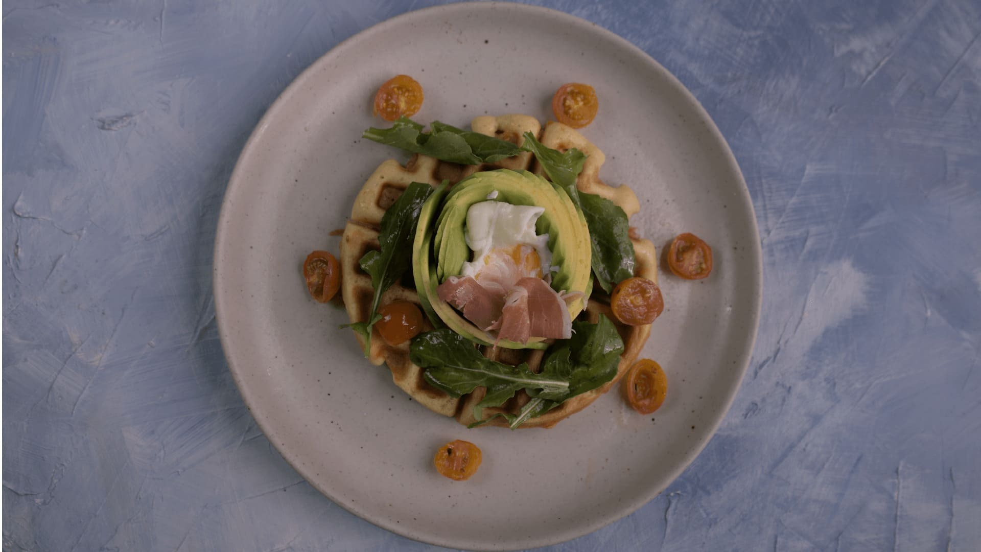 Un gofre salado con aguacate, huevo pochado, jamón serrano y tomates cherry, en un plato blanco.