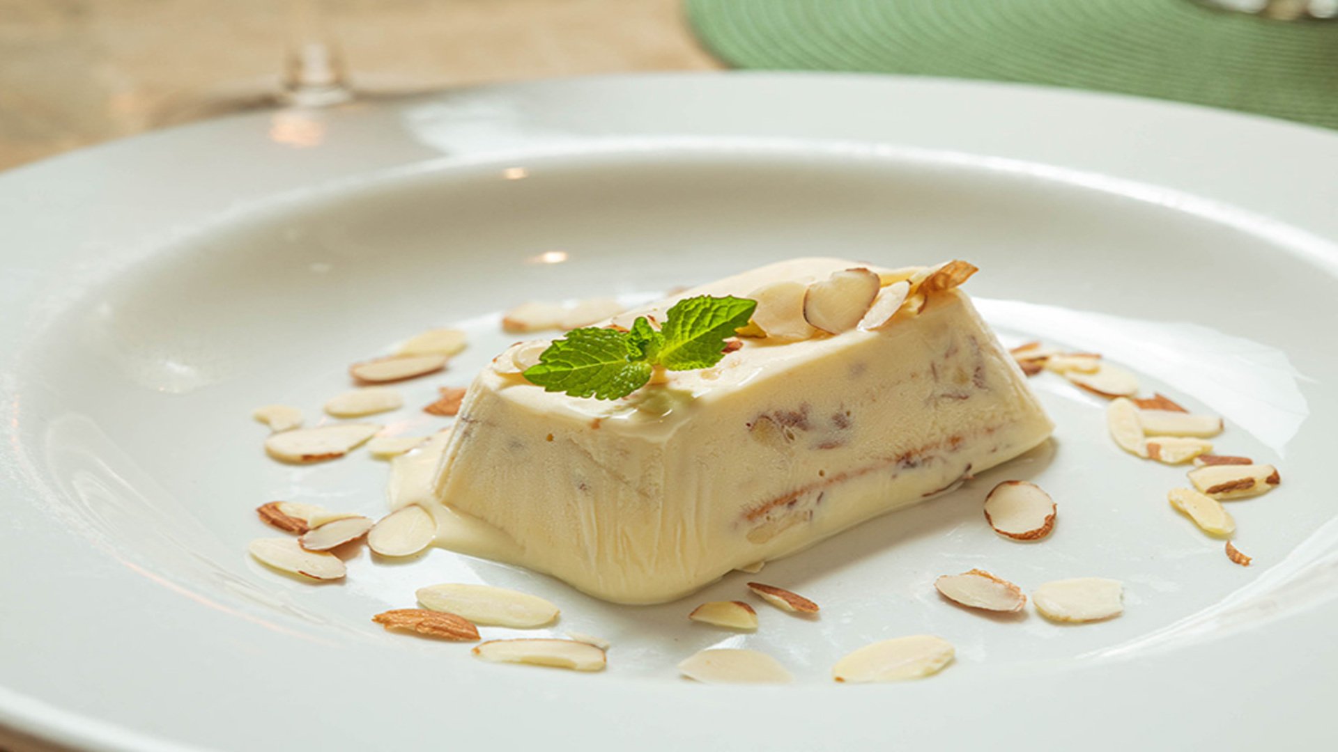 Un postre helado tipo parfait con almendras laminadas y una hoja de menta en un plato blanco.