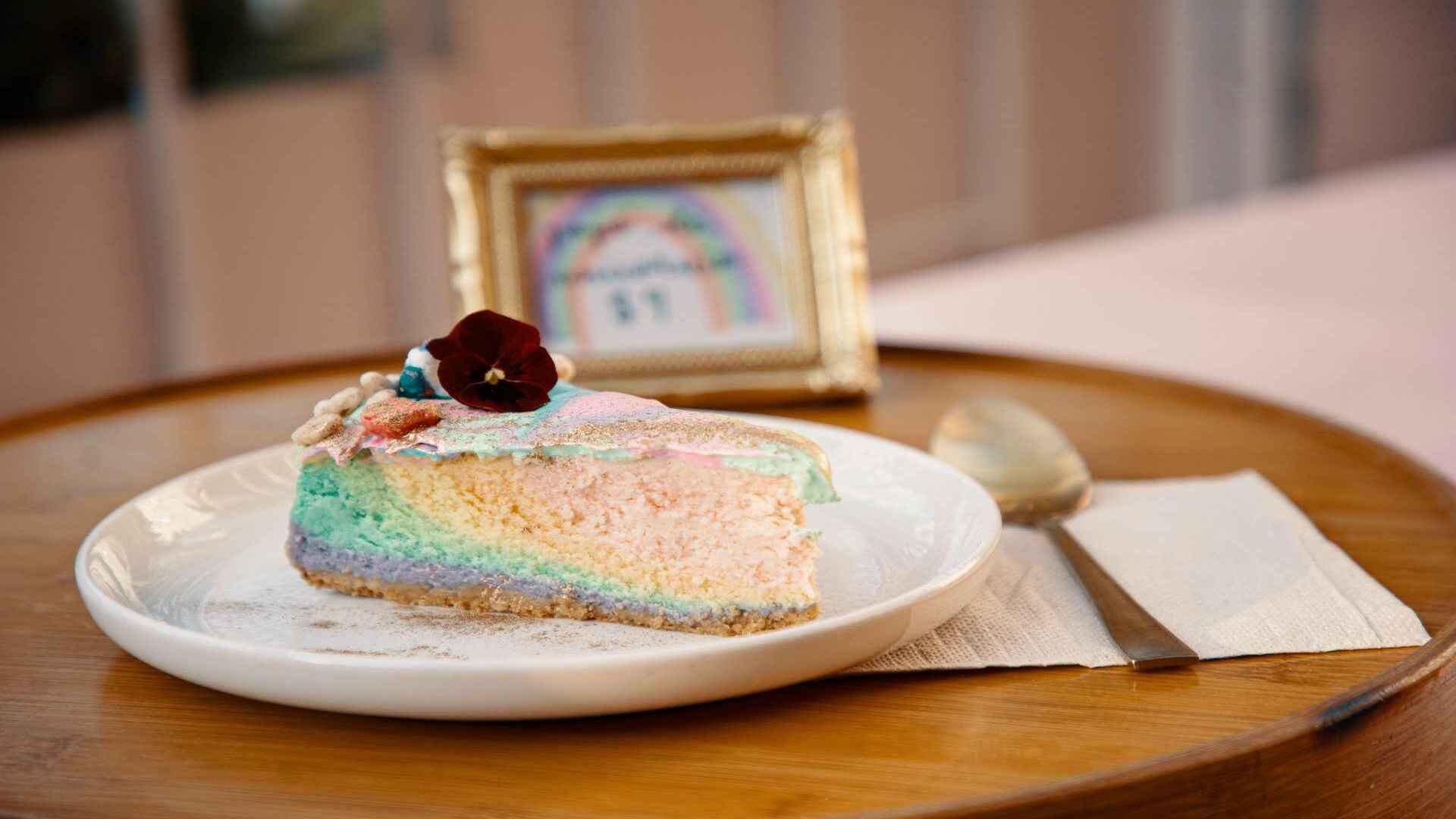  Una rebanada de pastel arcoíris con glaseado brillante y una flor en la parte superior, servida en un plato blanco.