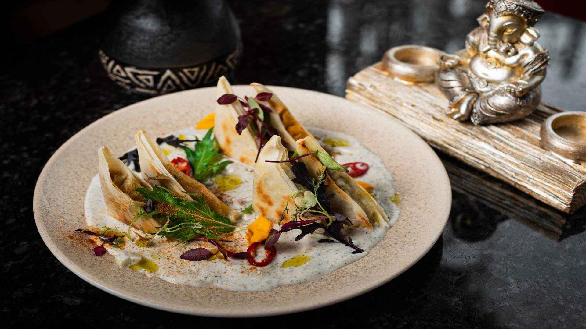 Empanadillas con hierbas, chiles y pétalos sobre una salsa blanca cremosa, con una estatua de Ganesha.