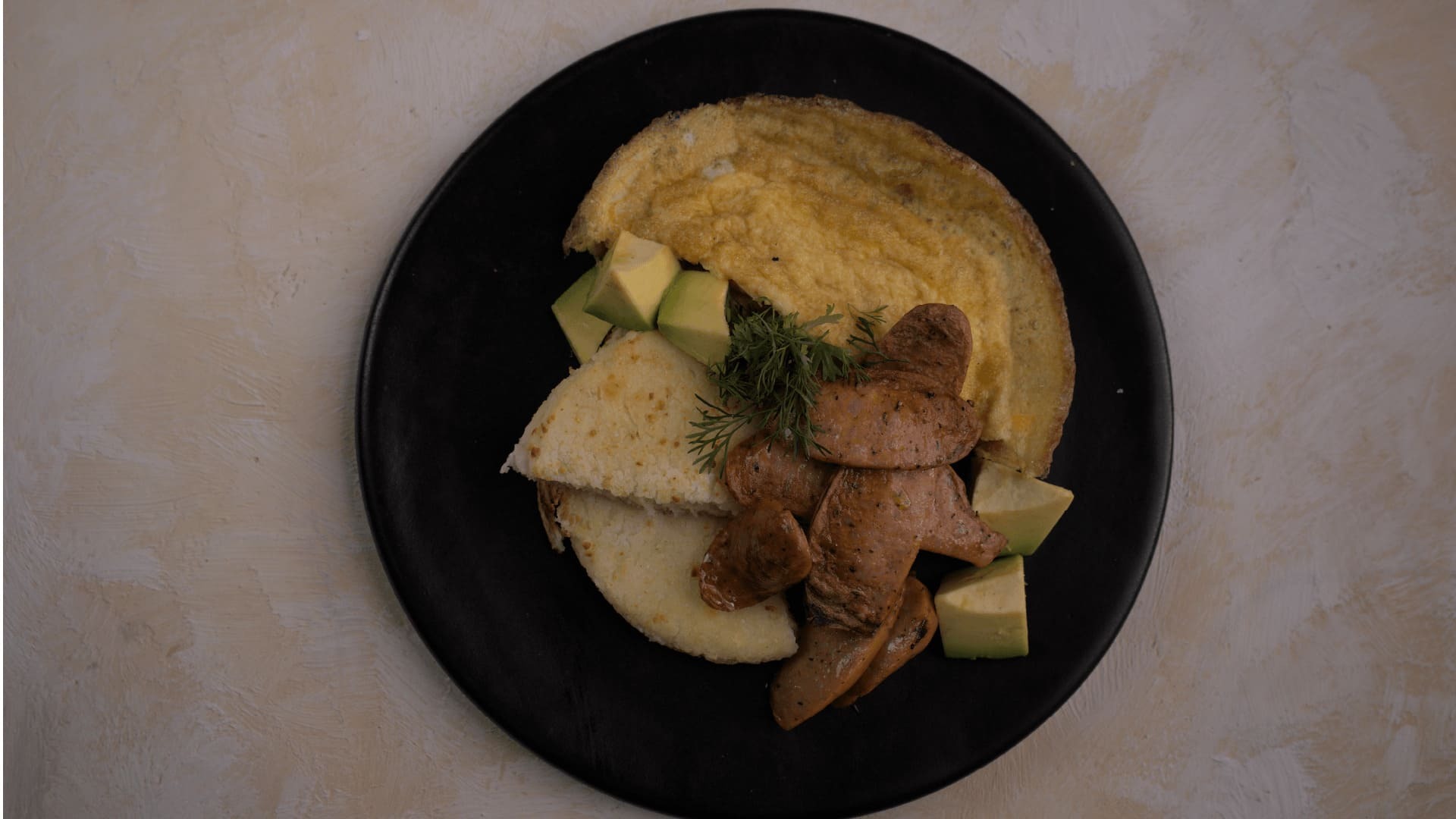 Un plato de desayuno con omelette, aguacate, arepa y salchichas, servido en un plato negro.