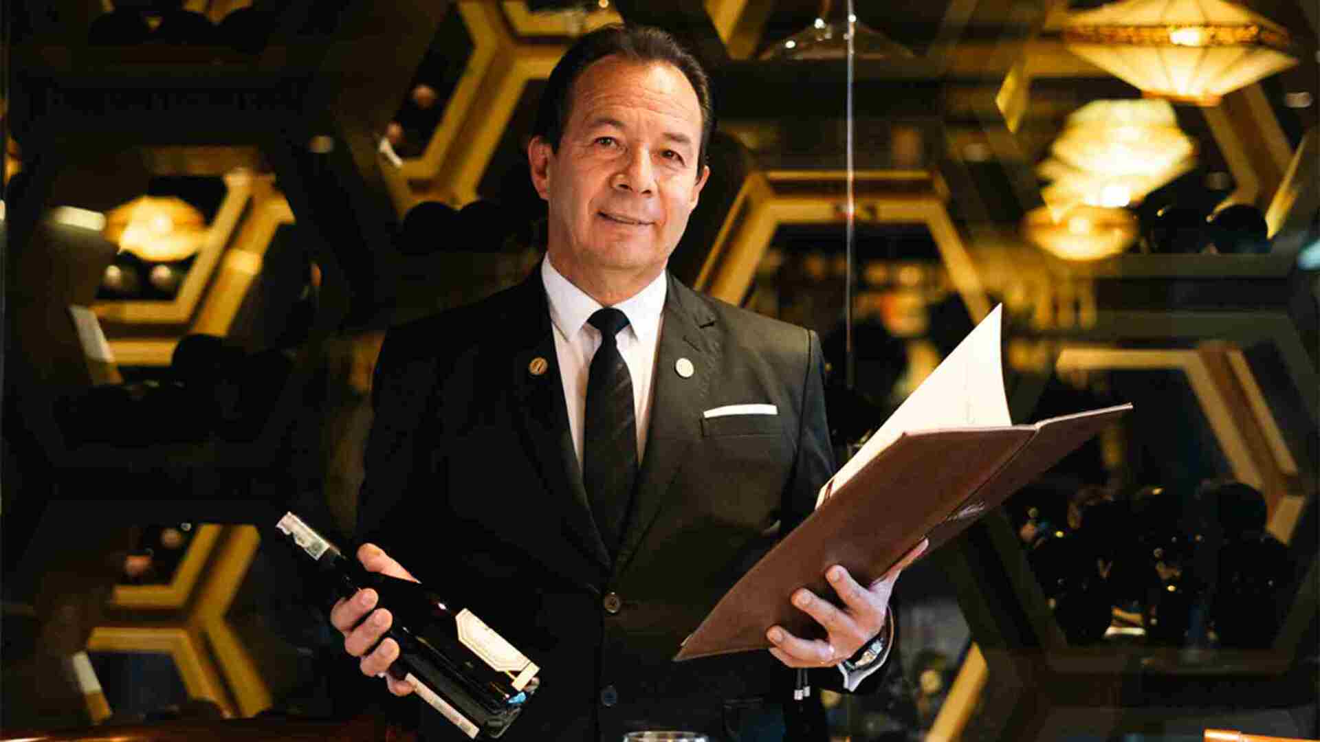 Un sommelier sonriente, vestido elegantemente, sostiene una botella de vino y una carta de bebidas en un restaurante.