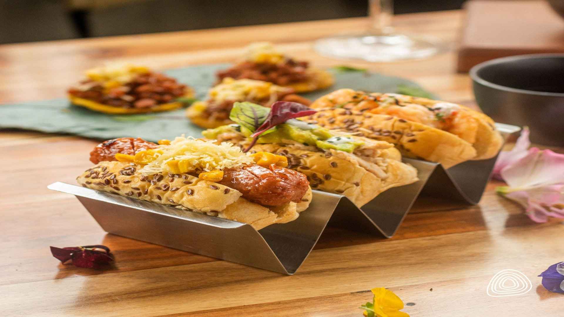 Tres mini hot dogs gourmet con aguacate, queso y semillas de sésamo, en un soporte metálico.