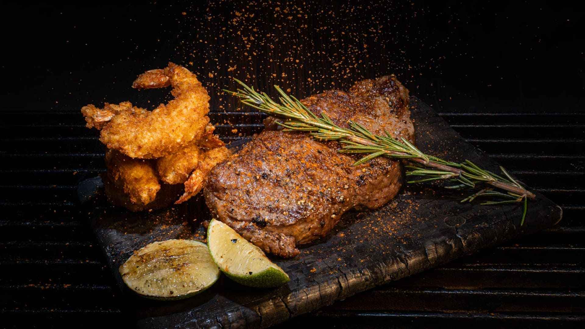 Un jugoso filete con camarones empanizados, romero y rodajas de lima, servido en una tabla de madera.