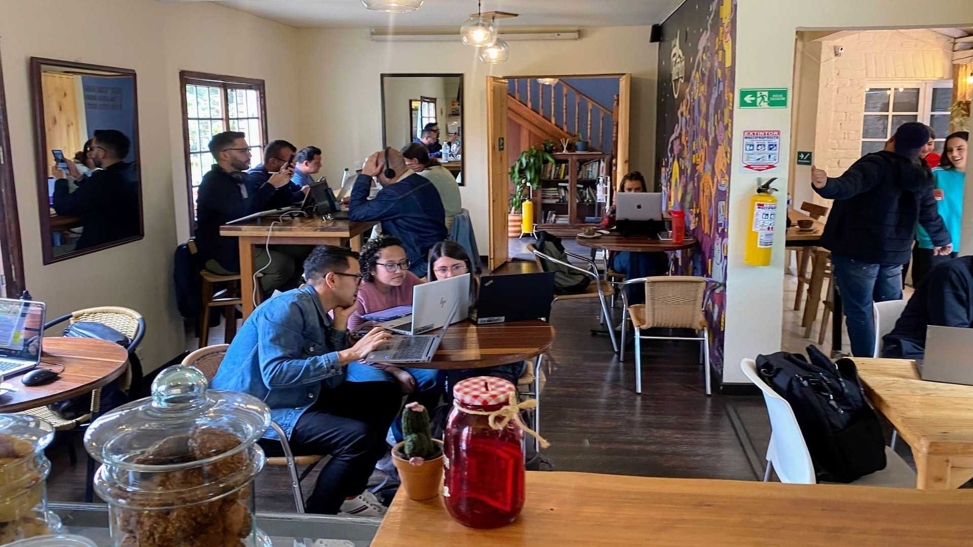 Interior de una cafetería con gente trabajando en laptops, arte mural y un ambiente relajado y productivo.