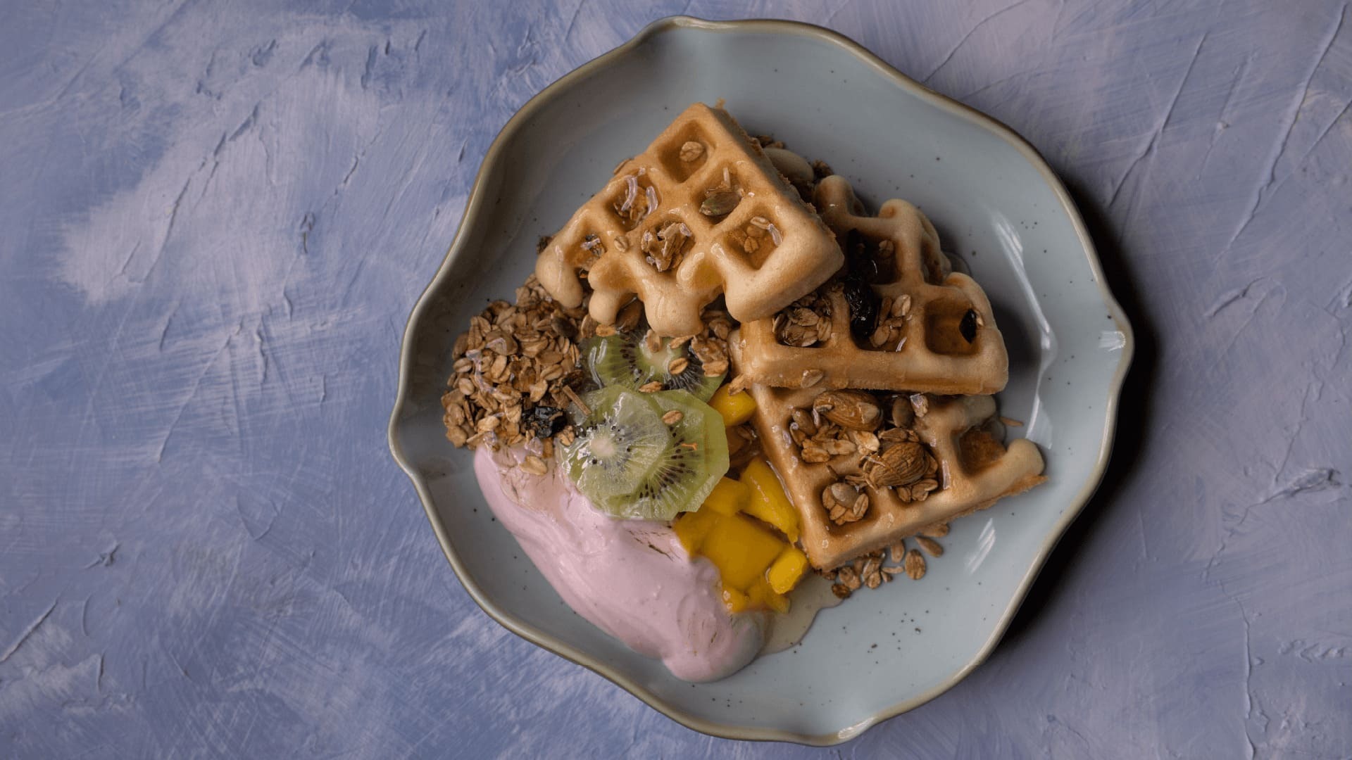 Gofres con yogur rosa, granola, kiwi y mango, en un plato ondulado.