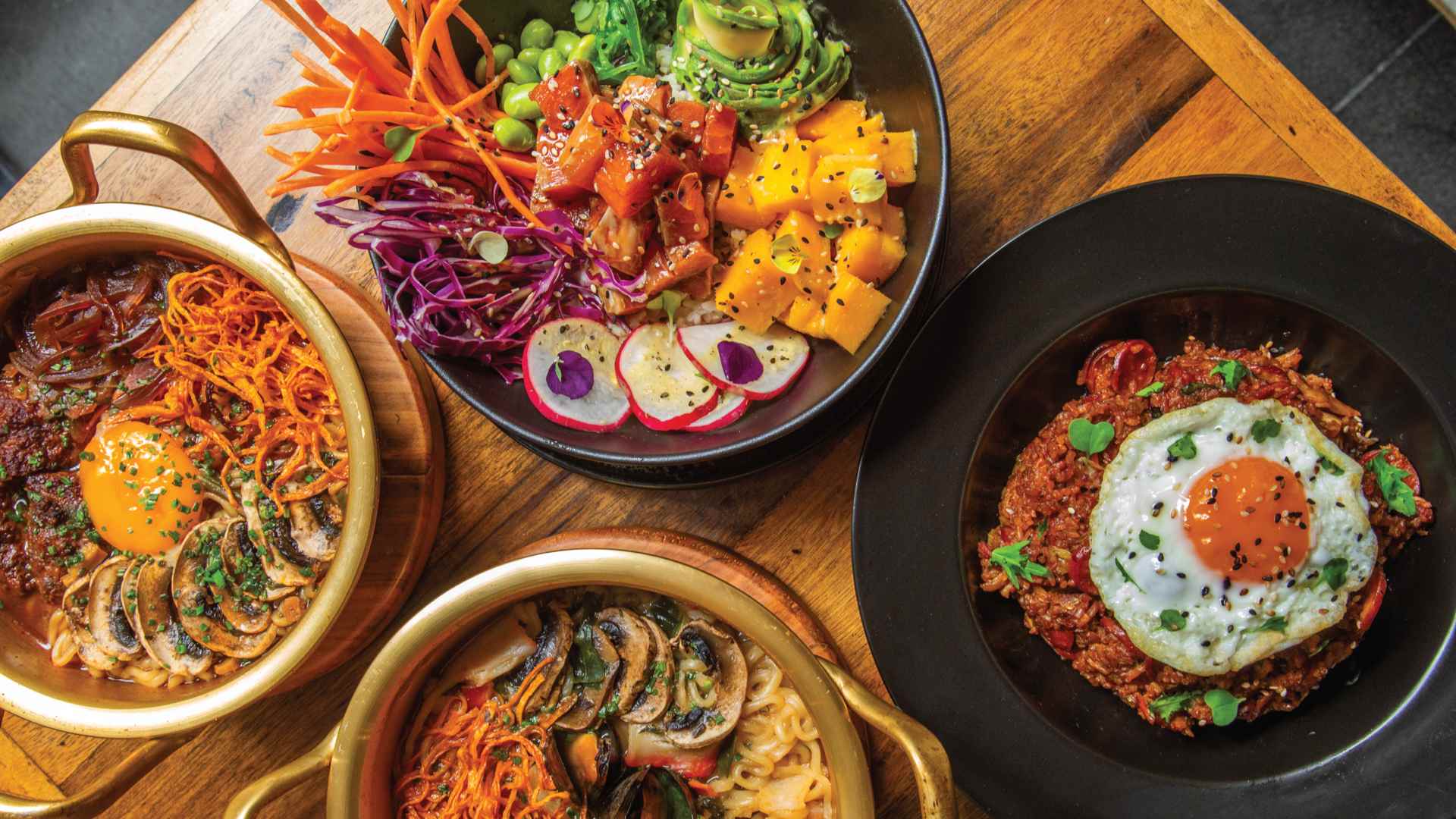 Una colorida y abundante mesa con un poke bowl, ramen con huevo y un plato de arroz frito, mostrando una fusión asiática.