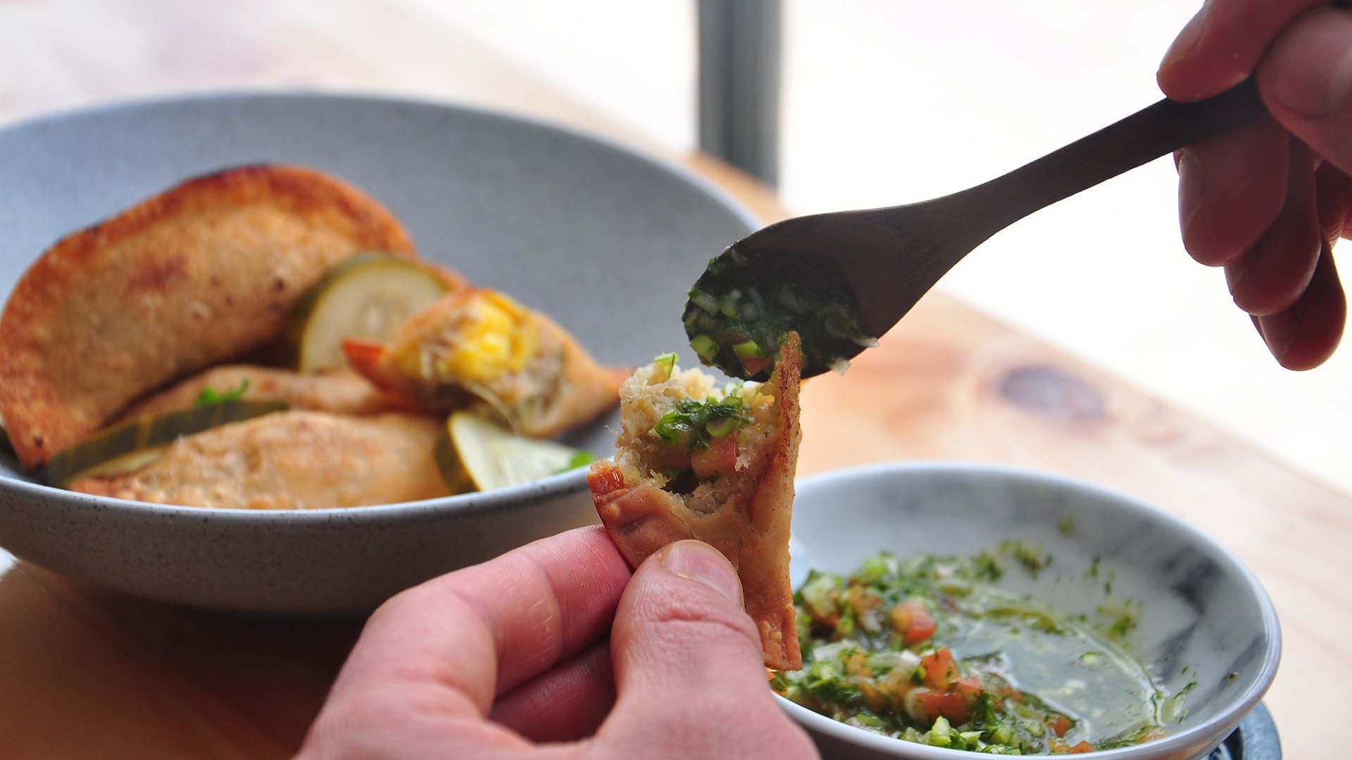 Manos sirviendo salsa chimichurri sobre una empanada frita, con un plato de empanadas al fondo.