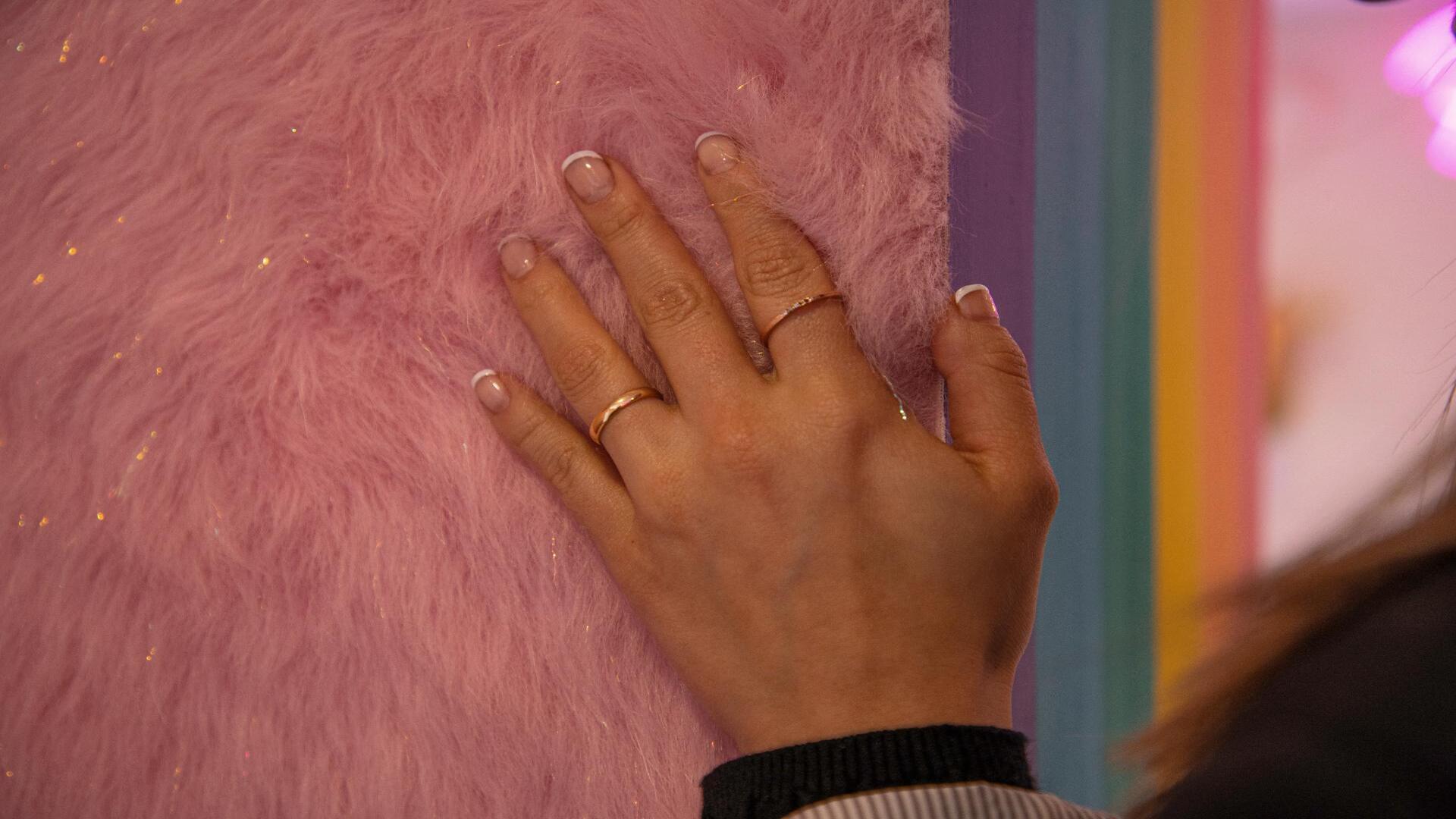 Una mano con anillos dorados y manicura francesa toca suavemente una superficie de textura peluda y rosa brillante.