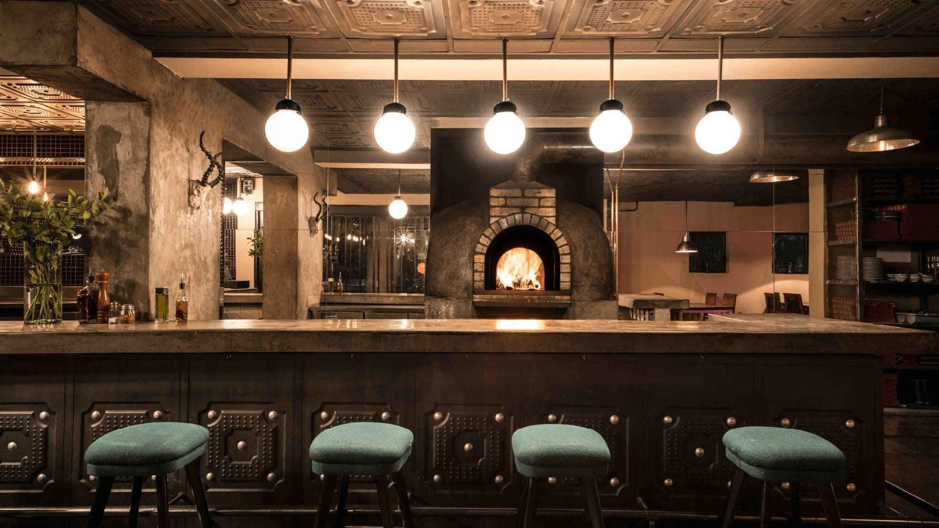 Un bar con horno de ladrillo, taburetes verdes y luces colgantes, creando un ambiente rústico.