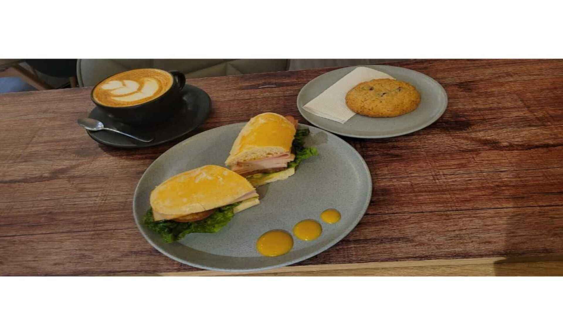 Un café latte con un sándwich recién hecho y una galleta con chispas de chocolate, sobre una mesa de madera.