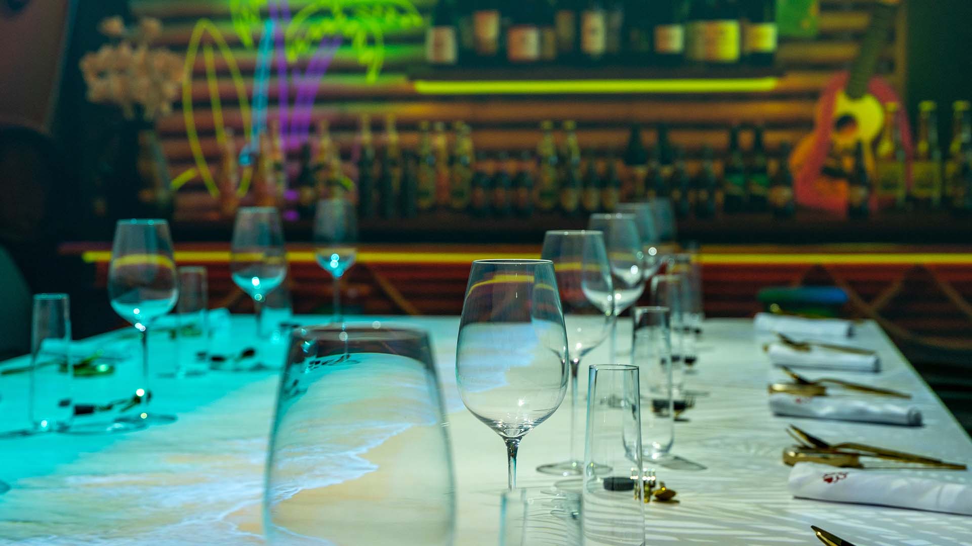 Mesa elegantemente puesta con copas vacías, lista para una noche especial con proyecciones vibrantes de un bar.