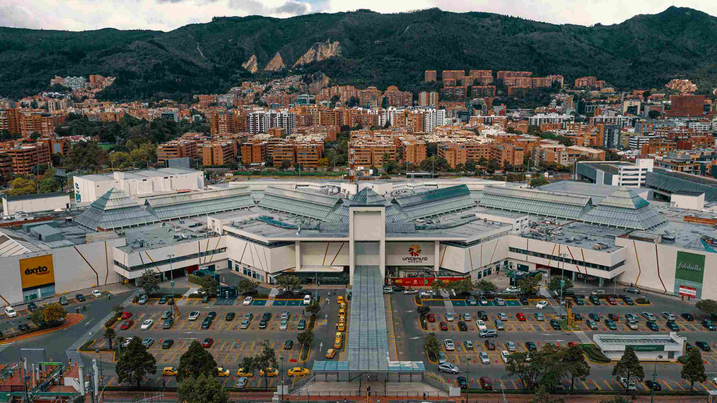  Vista desde arriba de la edificación del Centro Comercial Unicentro con su moderna estructura en concreto de color blanco, una amplia zona de parqueaderos. Al fondo se ven edificios que llegan hasta en los cerros orientales.