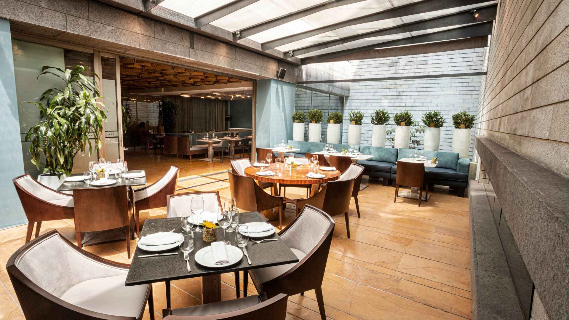 Un restaurante moderno con una sección interior y una terraza cubierta, mesas elegantes y cómodos sofás. La decoración es sofisticada, con plantas y tonos neutros.
