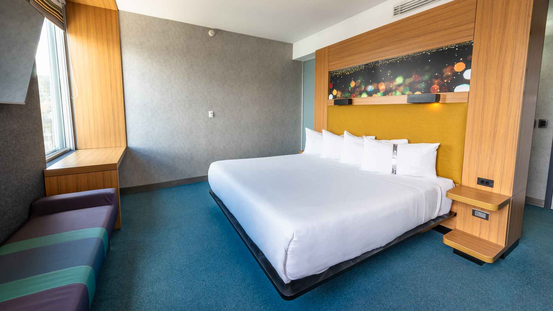 Una habitación de hotel Aloft moderna y colorida con una cama king-size, una ventana con banco y un cabecero decorativo con luces.
