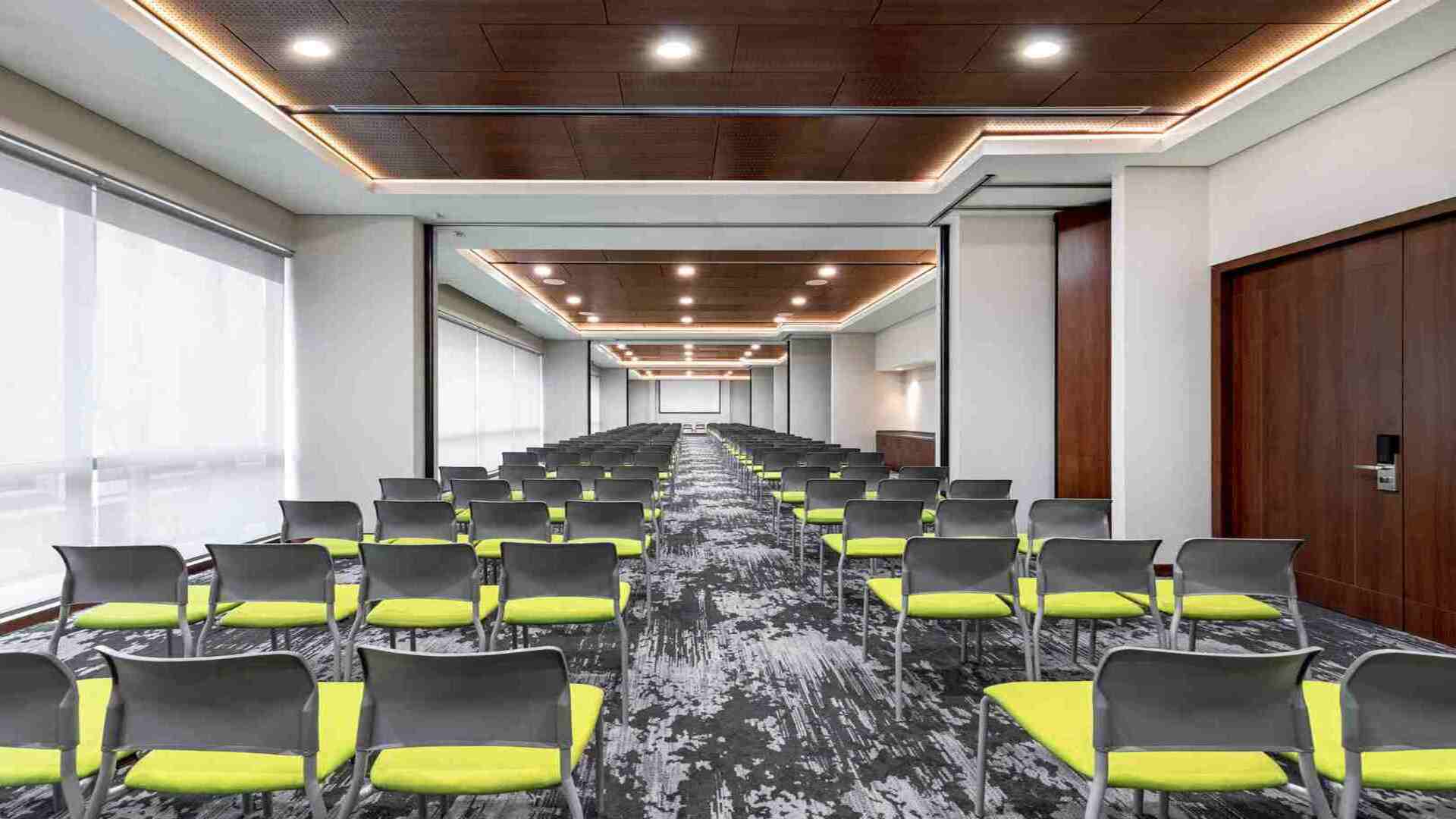 Un moderno lobby lounge con sofás verdes y grises, mesas de madera y grandes lámparas colgantes. La decoración cuenta con paneles de madera y patrones geométricos.