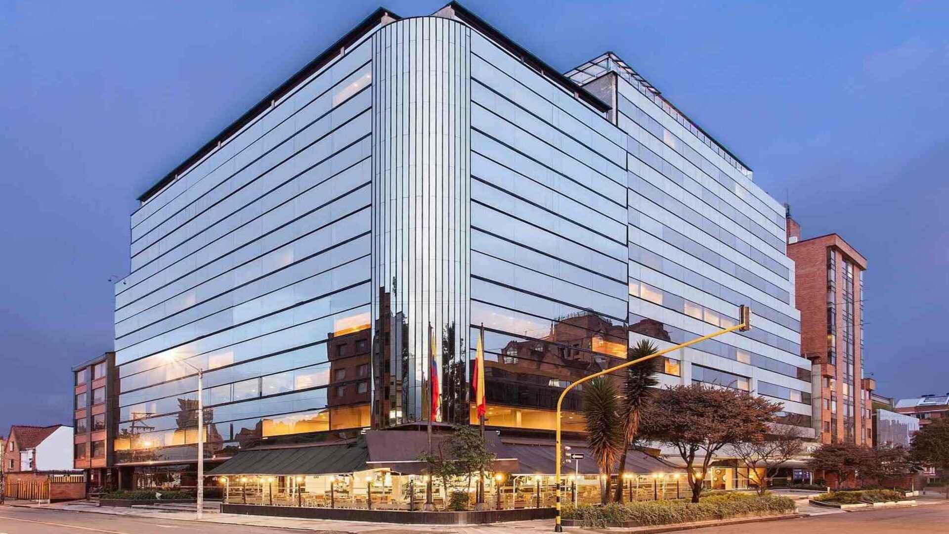  Un moderno edificio de hotel con fachada de cristal, que refleja el entorno urbano al atardecer o al anochecer. La iluminación del restaurante en la planta baja añade calidez.
