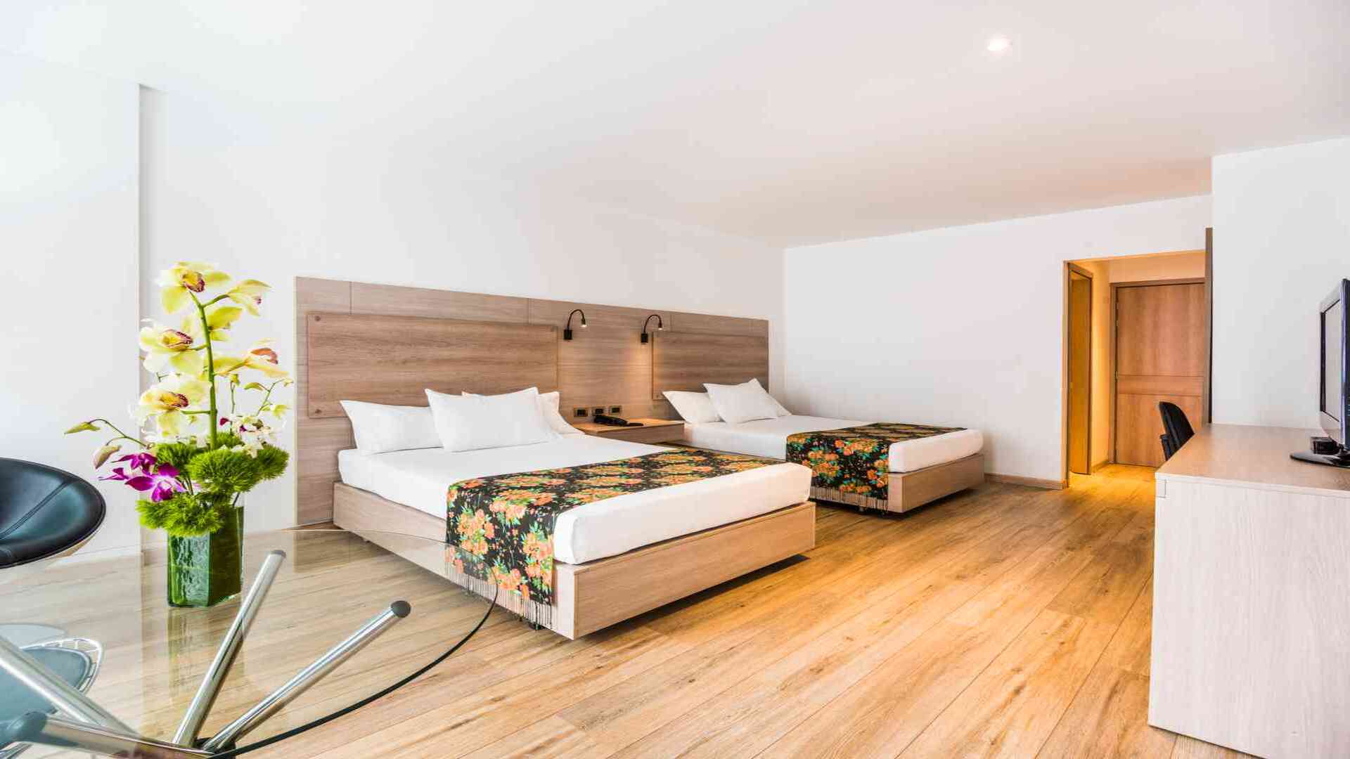 Una habitación de hotel luminosa y moderna con dos camas individuales, cabeceras de madera clara y caminos de cama con motivos florales. Orquídeas añaden un toque de color.
