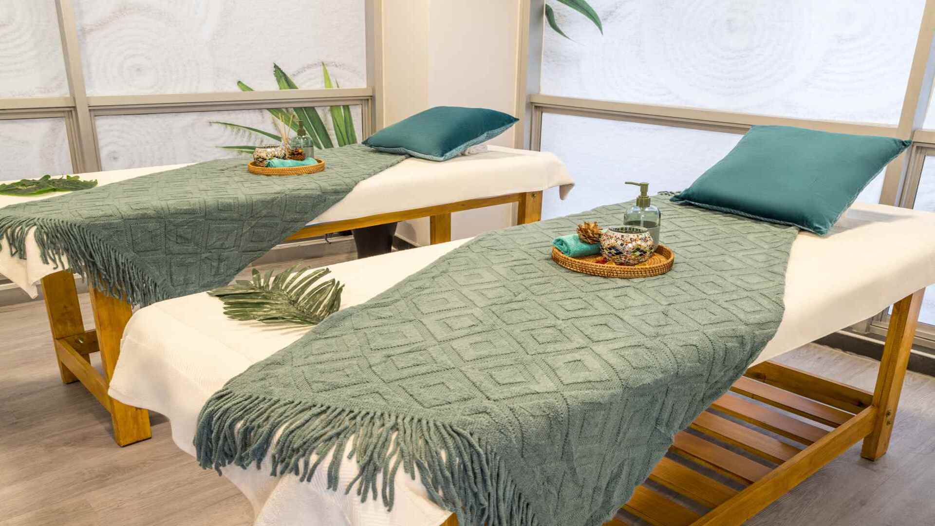 Una sala de spa con dos camillas de masaje preparadas, con mantas y almohadas en tonos verdes. El ambiente es tranquilo y relajante.