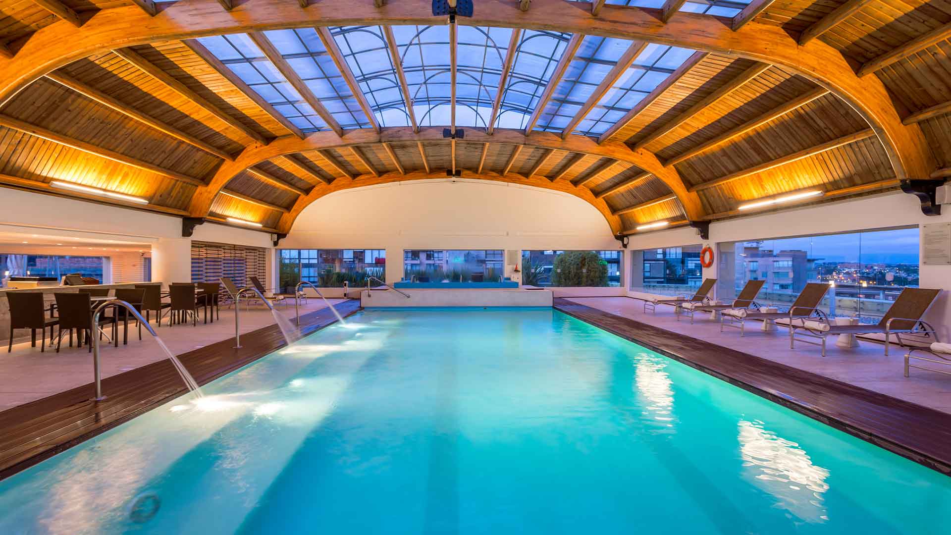 Piscina: Piscina cubierta con techo de madera arqueado, chorros de agua y tumbonas junto a ventanales. Vista urbana y luz cálida.