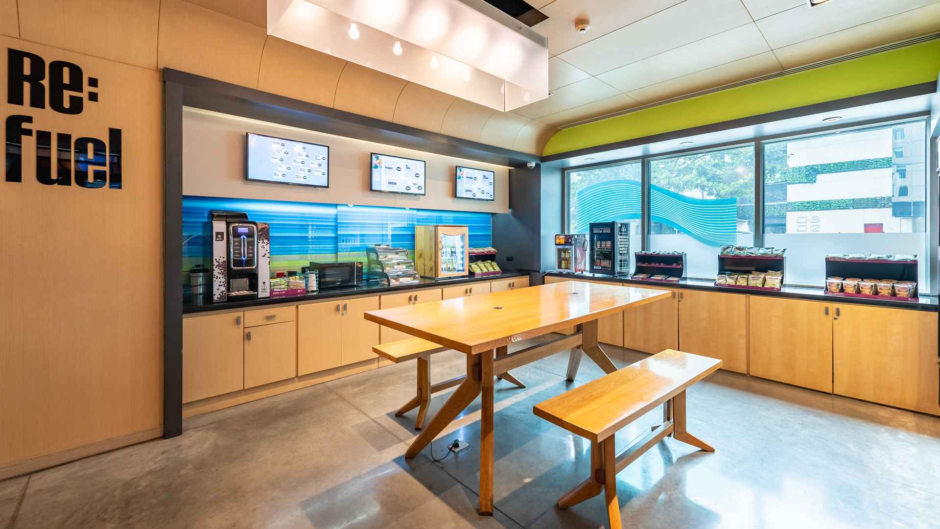 Una estación de autoservicio "Re:fuel" con opciones de café, bebidas y snacks, mesas comunitarias de madera y grandes ventanales.