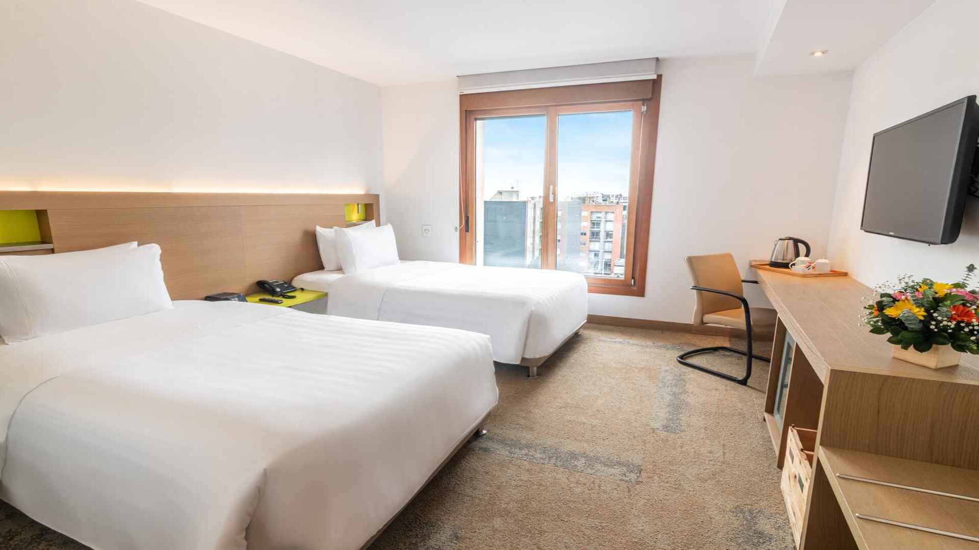 Una luminosa habitación doble con dos camas individuales, un escritorio con silla y una televisión montada en la pared. Grandes ventanas dejan entrar mucha luz natural.