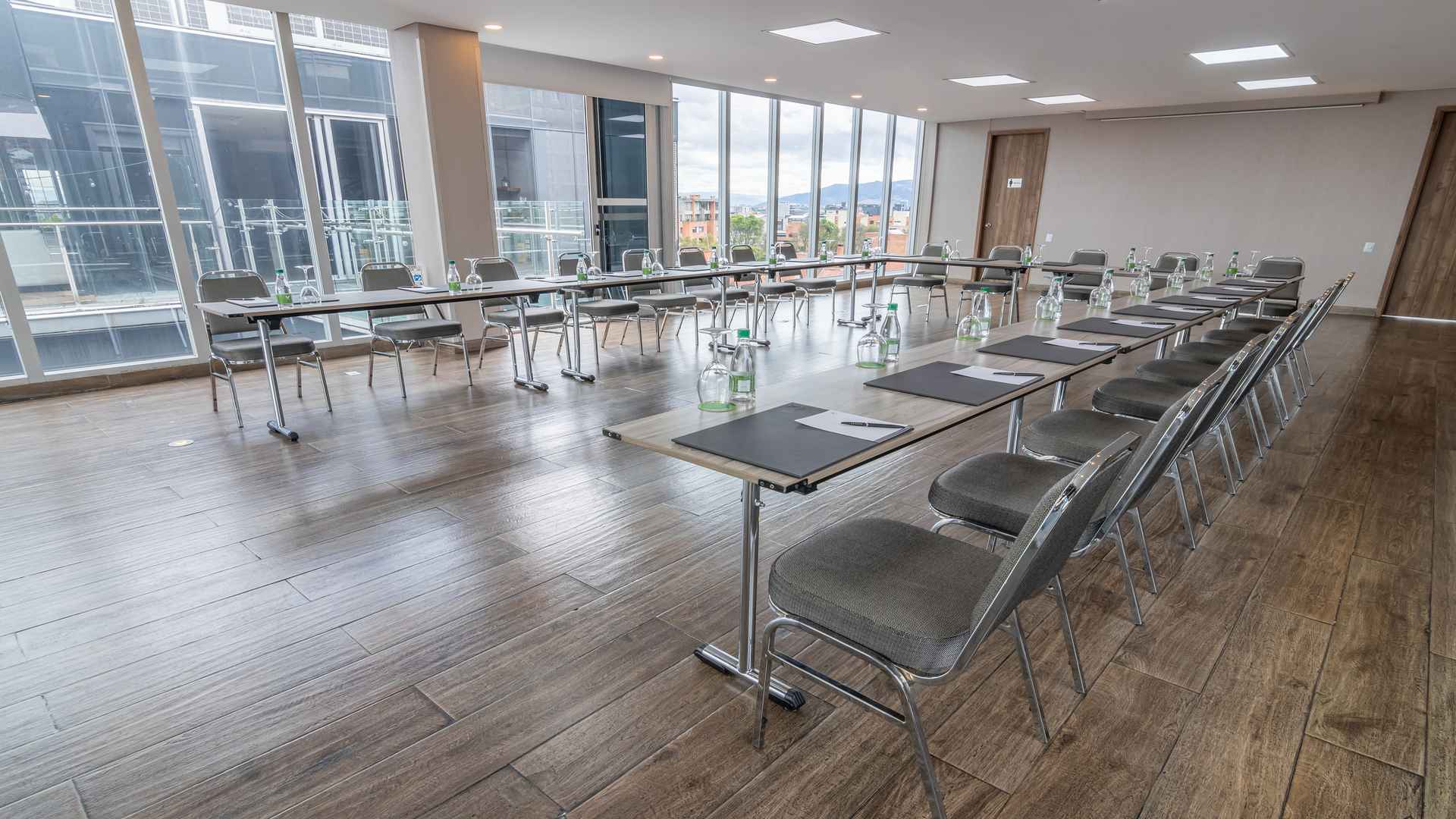  Una sala de reuniones moderna con mesas dispuestas en un gran rectángulo, sillas grises y grandes ventanales que ofrecen vistas urbanas.