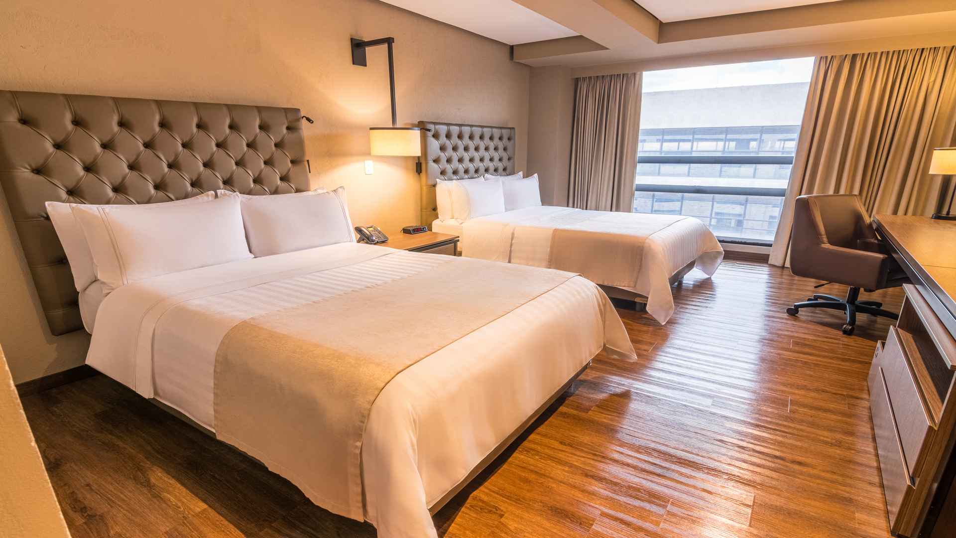Habitación de hotel con dos camas dobles, cabeceras capitoné y ventanas grandes.