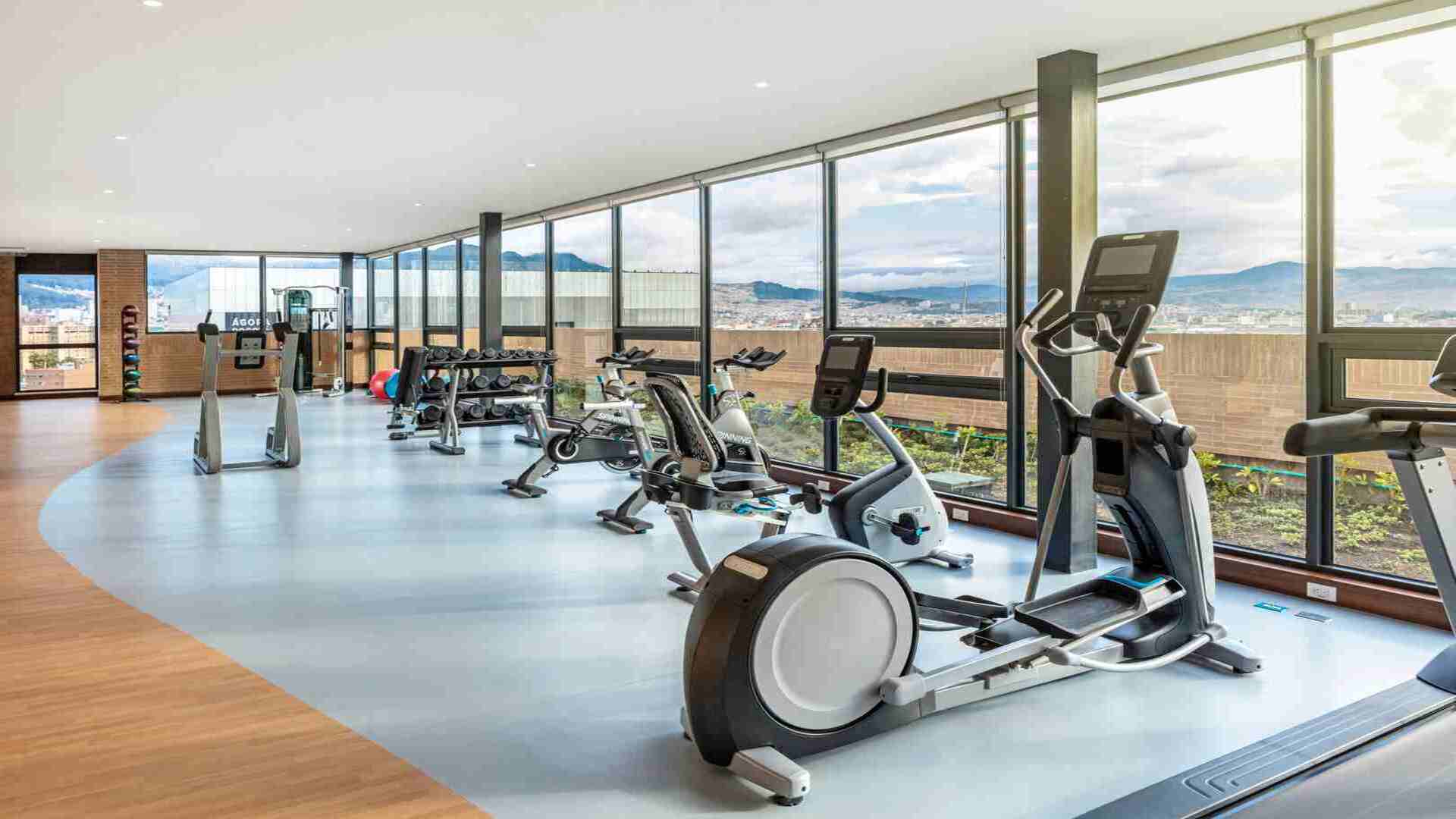 Un gimnasio espacioso y bien iluminado con grandes ventanales que ofrecen vistas panorámicas de la ciudad y las montañas. Equipado con diversas máquinas de ejercicio.