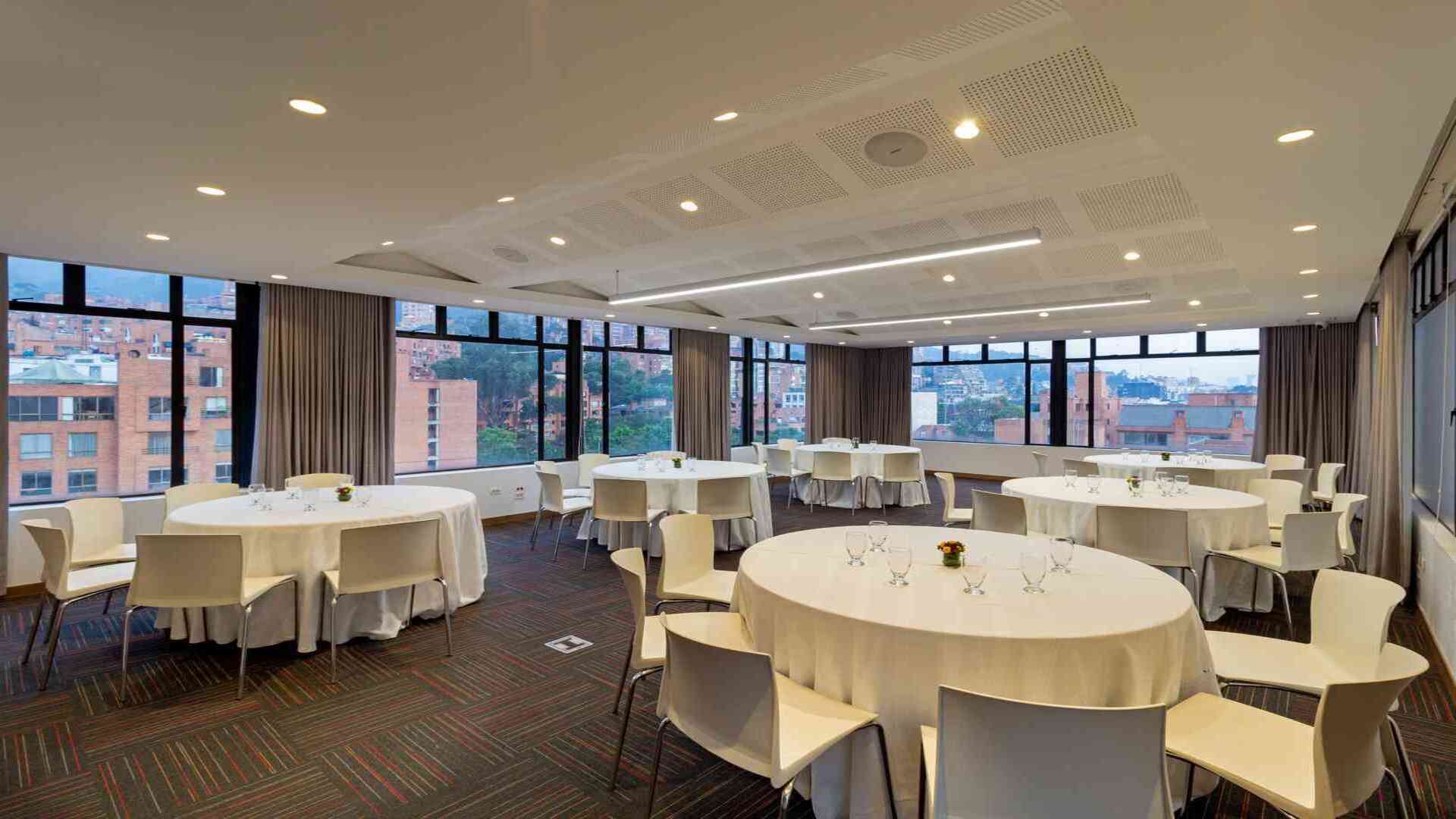 Una sala de eventos dispuesta con mesas redondas vestidas de blanco y sillas, ideal para banquetes o conferencias. Grandes ventanales ofrecen vistas a la ciudad.