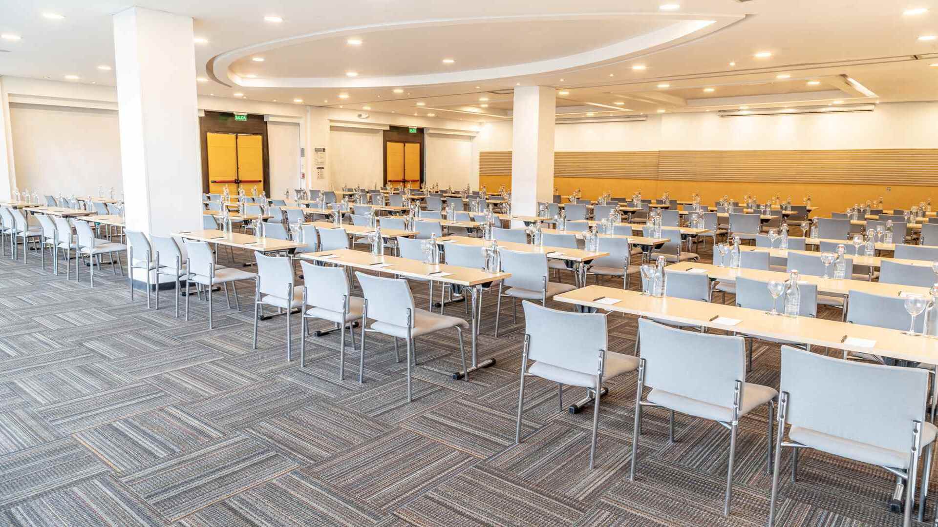Una gran sala de conferencias preparada en estilo aula, con numerosas mesas y sillas dispuestas ordenadamente. Ideal para eventos y capacitaciones.