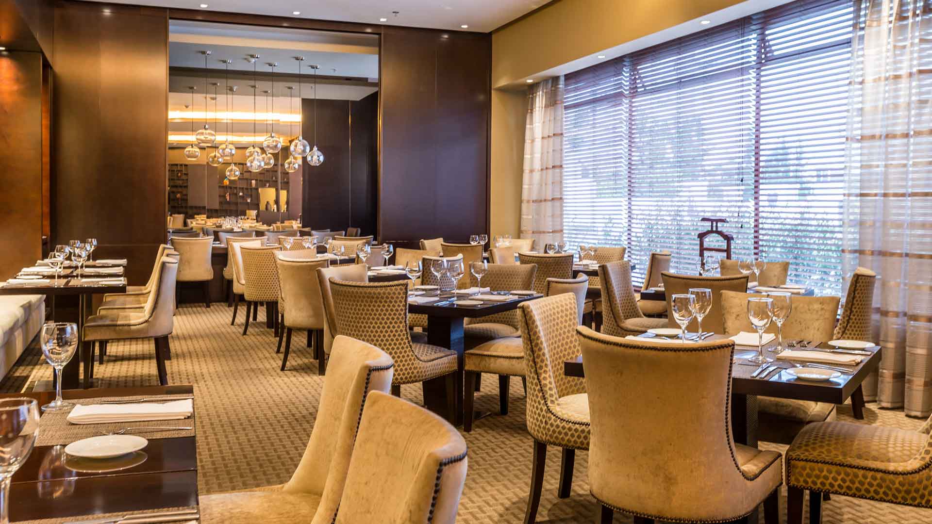 Restaurante: Restaurante moderno con sillas beige, mesas ordenadas, ventanales con persianas y lámparas. Ambiente y servicio amable.