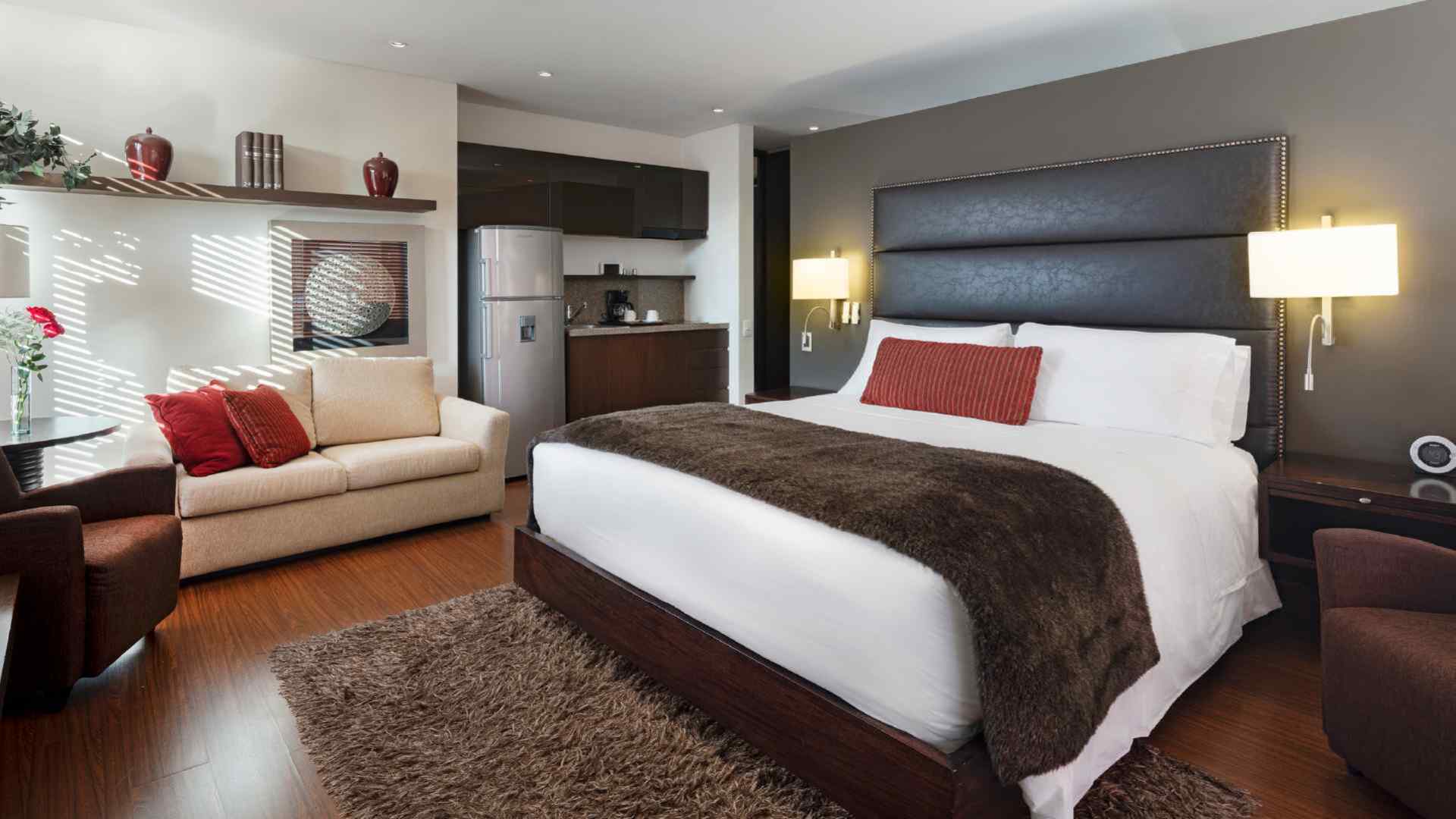 Una suite de hotel espaciosa y moderna con una cama king-size, sofá, alfombra de pelo y una pequeña cocina integrada. La decoración es elegante y funcional.