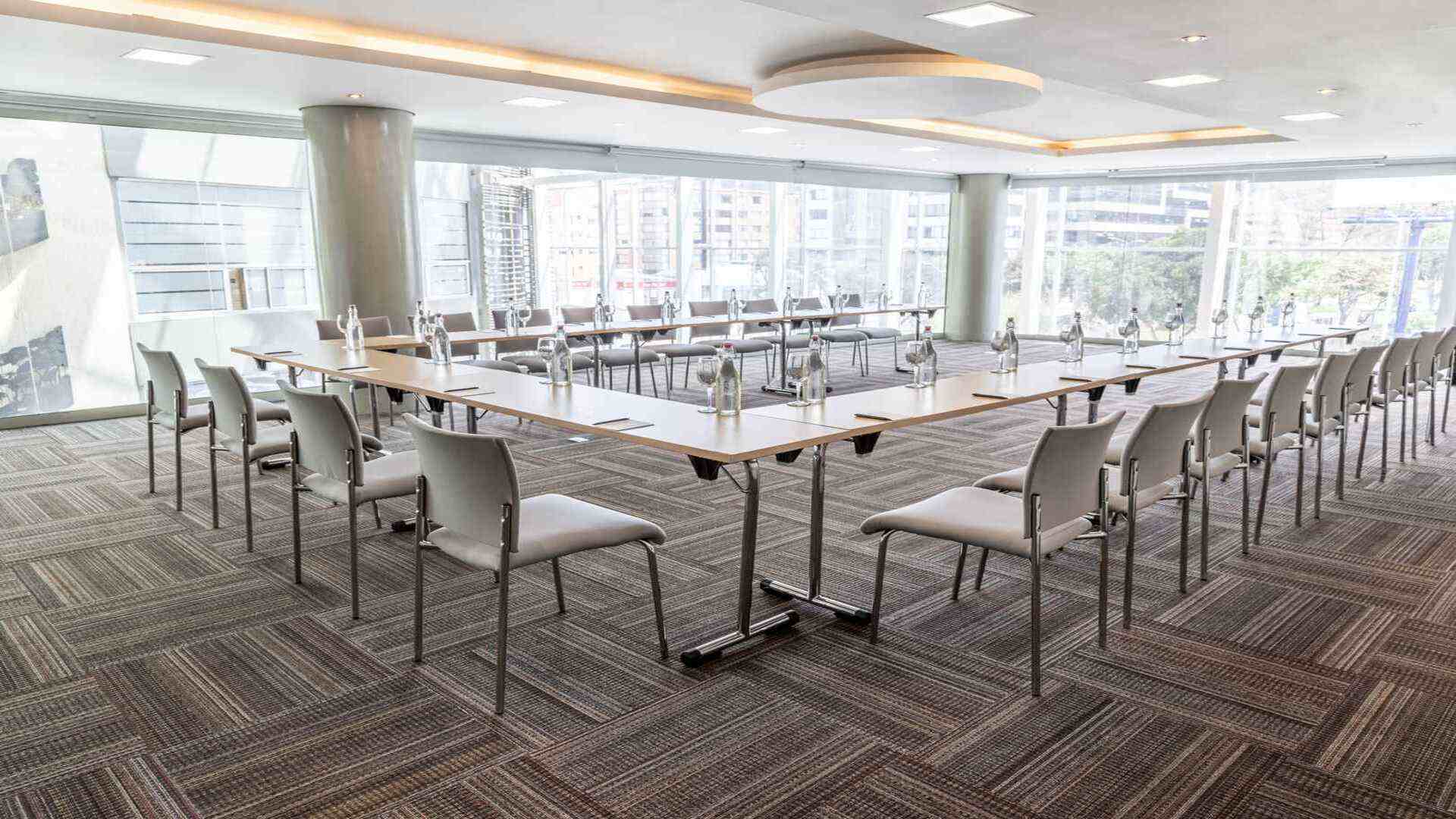  Una sala de reuniones dispuesta en forma de "U" con mesas y sillas. Grandes ventanales ofrecen vistas de la ciudad, creando un ambiente de trabajo dinámico.