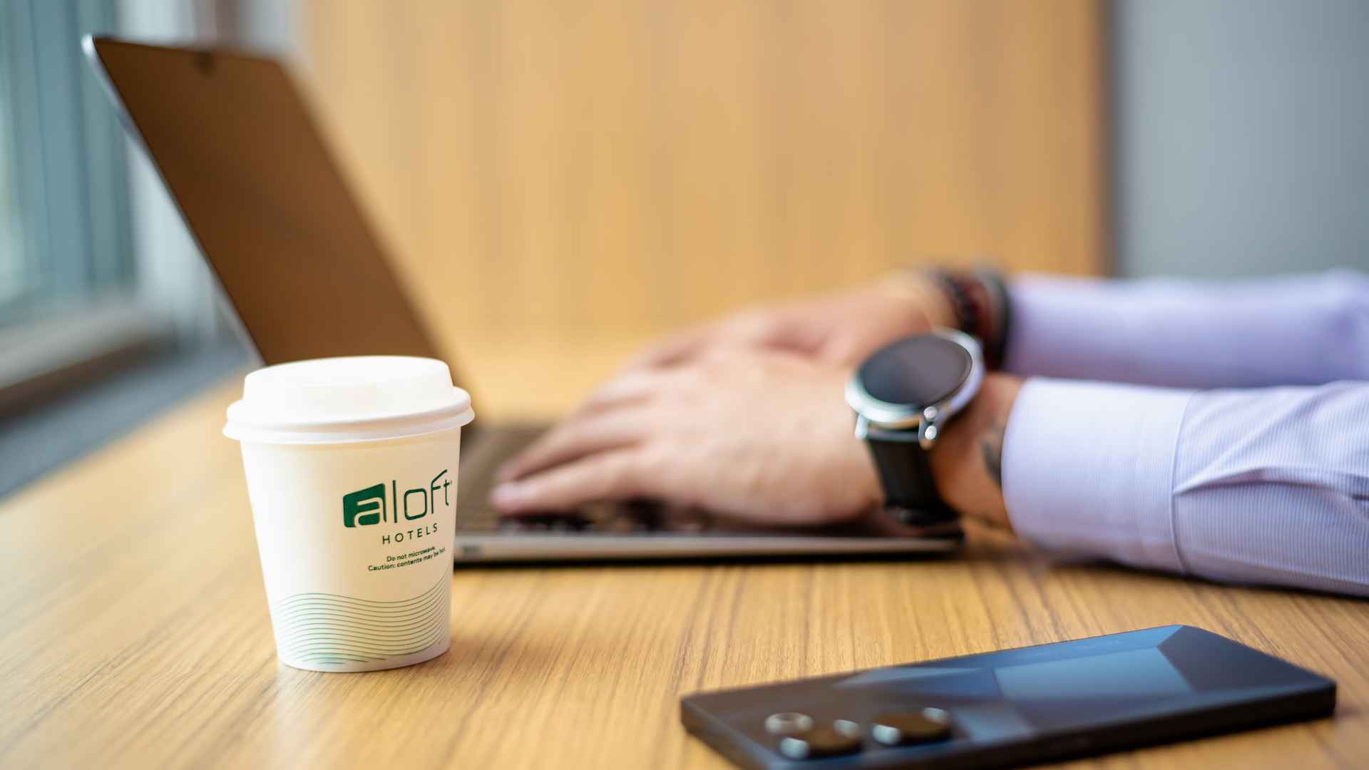 Un primer plano de una taza de café "Aloft Hotels" en un escritorio, con las manos de una persona trabajando en un portátil y un smartphone de fondo.