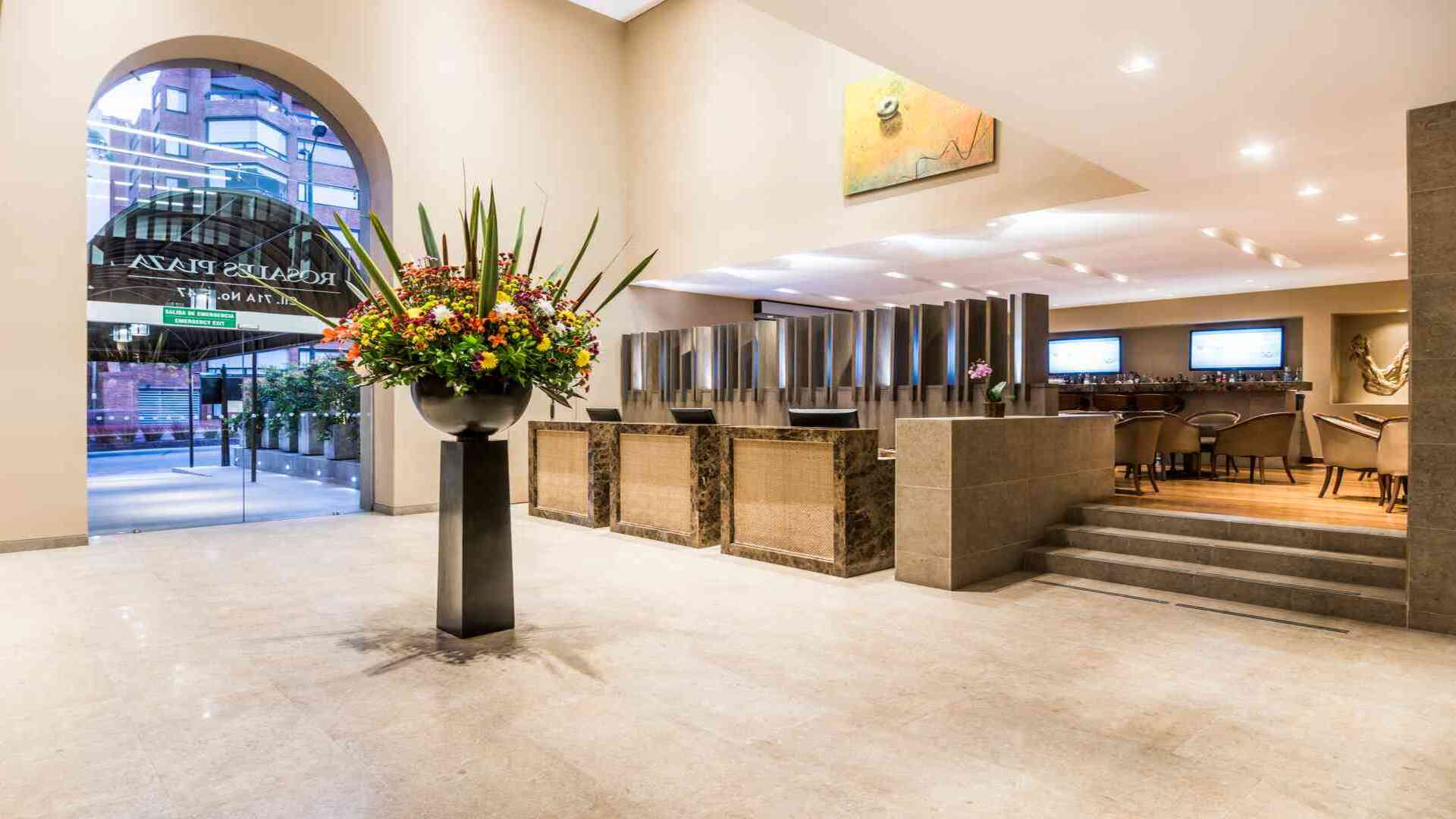El amplio y lujoso lobby de un hotel, con una gran composición floral, mostradores de recepción de mármol y un área de bar al fondo.