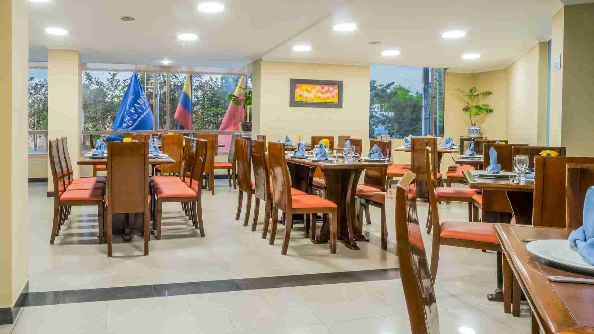  Un amplio restaurante de hotel con mesas de madera y sillas de cuero naranja, listo para servir a los comensales. Ventanas grandes permiten la entrada de luz natural.