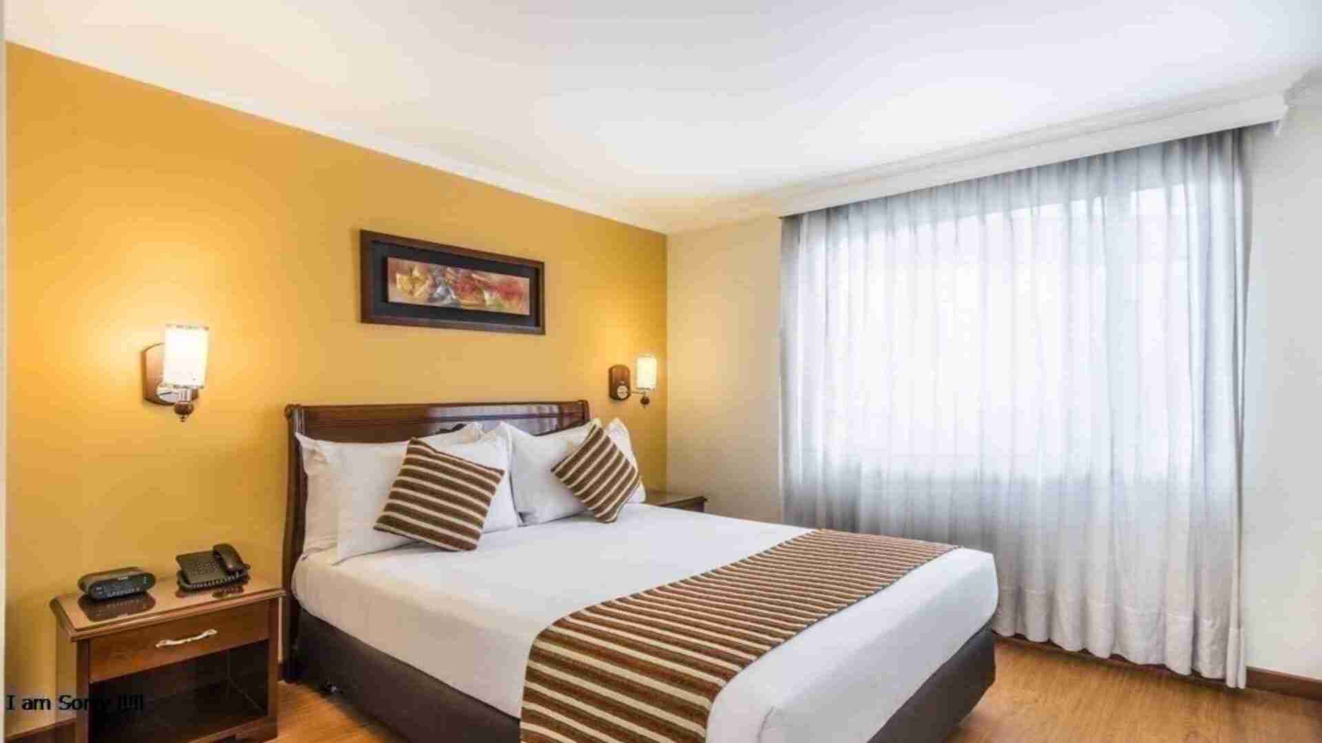 Un dormitorio de hotel con una cama doble, mesitas de noche y un televisor. La habitación es luminosa, con cortinas blancas y paredes en tonos amarillos.